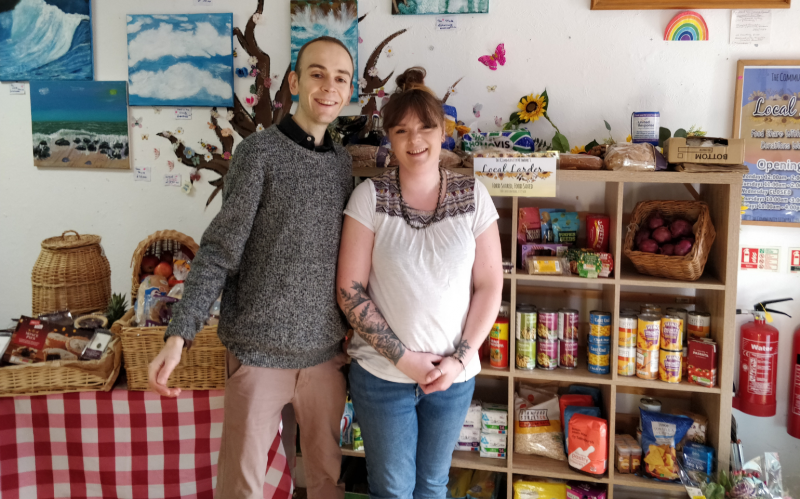 Oliver and Rachael stood in front of the larder. The shelves and baskets are full of tinned food, toiletries and fresh produce.