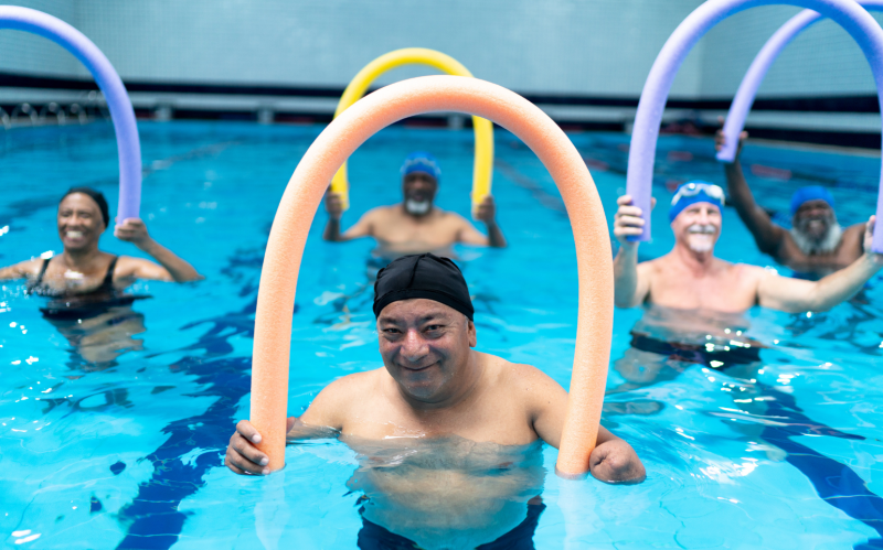 An older man is in an indoor swimming pool, taking part in a aqua fitness class or hydrotherapy session, surrounded by other people in the class.