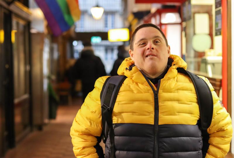 A man who has downs syndrome is wearing a yellow puffer jacket and a backpack is staring at the camera 
